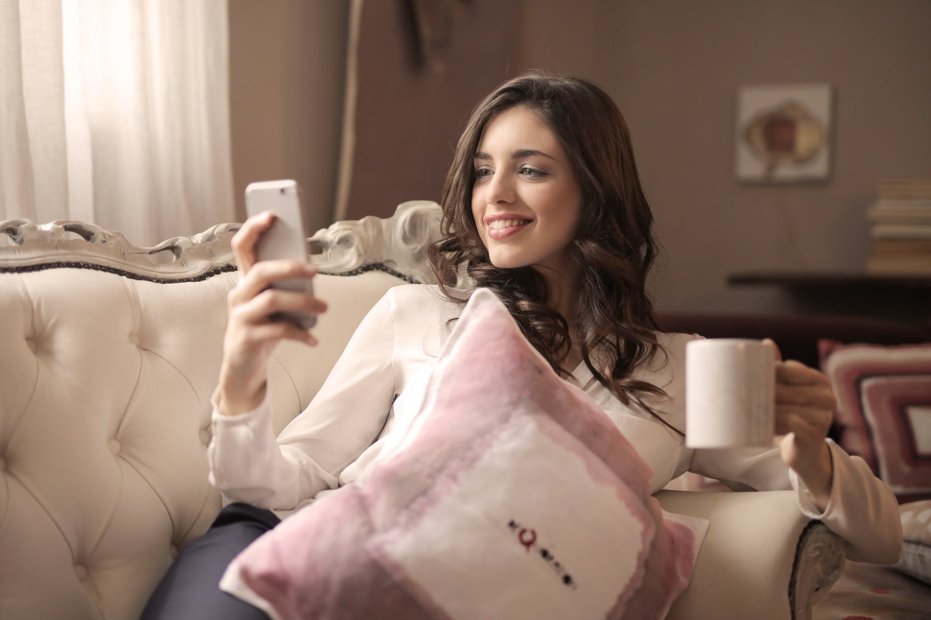 woman in white long sleeved shirt holding smartphone sitting on tufted sofa